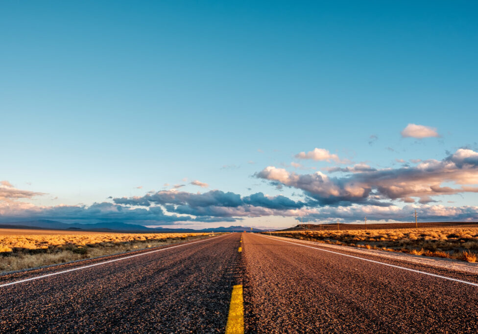 Image of an open highway leading towards the horizon meeting a bright blue sky with clouds in the distance